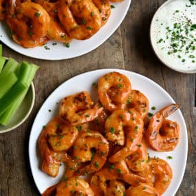 Grilled buffalo shrimp on a white plate next to a bowl with celery sticks and a bowl with ranch dressing.