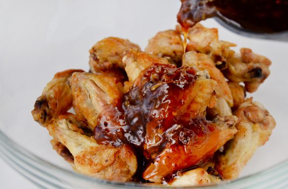 Baked chicken wings in glass bowl with orange sauce being poured over them.