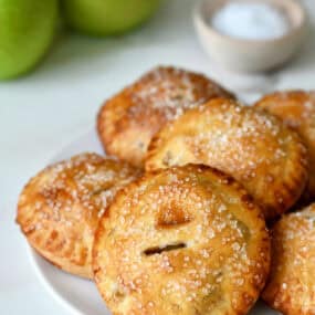 Salted caramel apple hand pies with sanding sugar on a white plate with Granny Smith apples in the background.