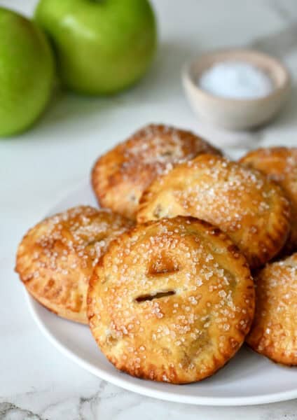 Salted caramel apple hand pies with sanding sugar on a white plate with Granny Smith apples in the background.