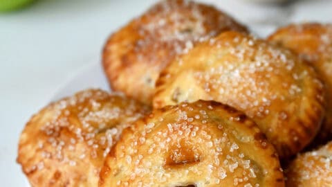 Salted caramel apple hand pies with sanding sugar on a white plate with Granny Smith apples in the background.