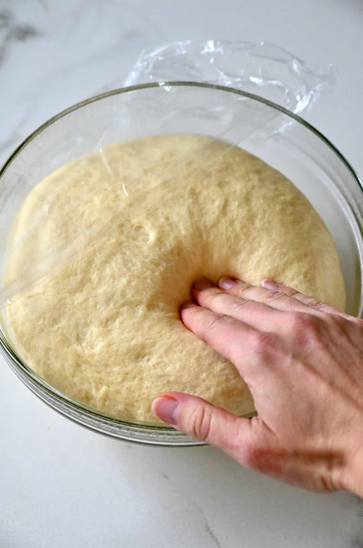 A hand presses into proofed dough in a glass bowl.