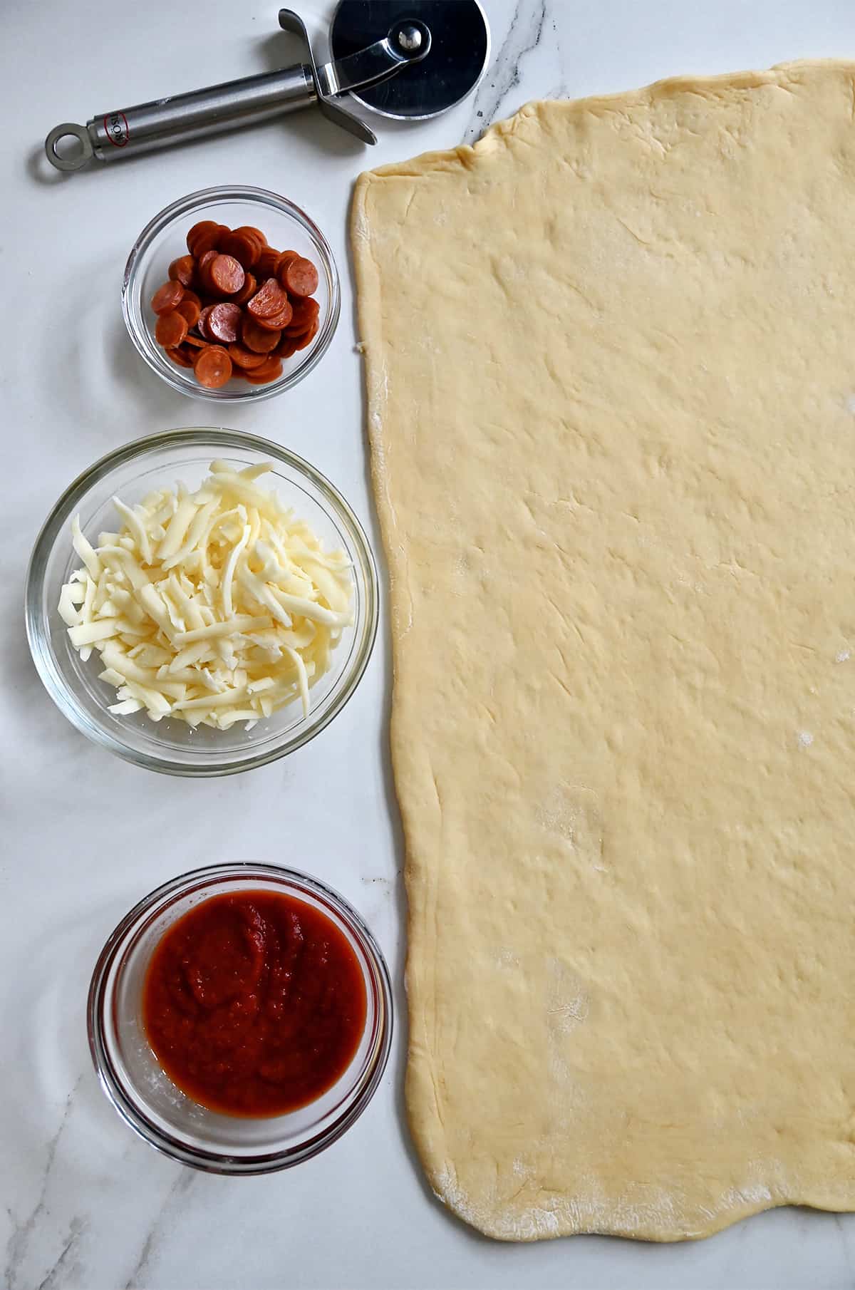Dough rolled out into a rectangle next to small bowls containing marinara sauce, shredded cheese and mini pepperonis.