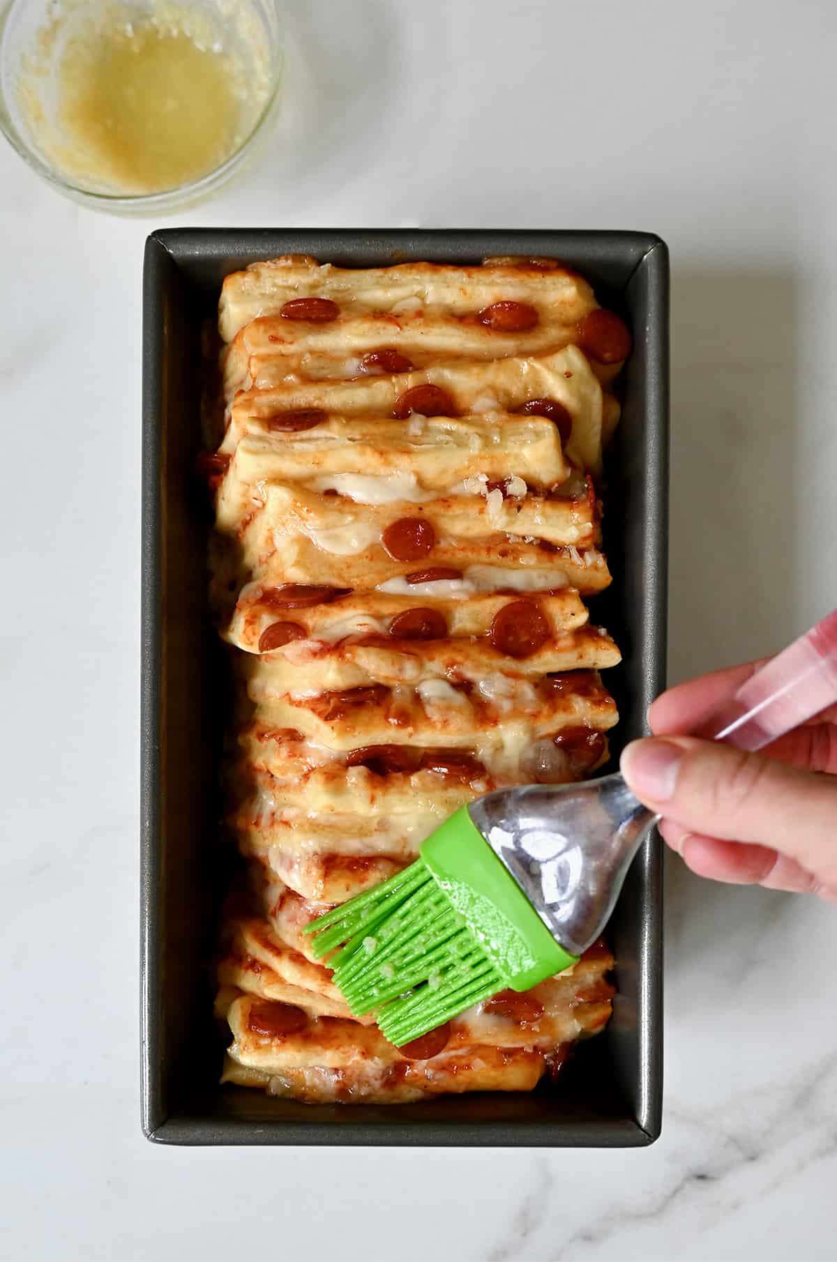 A hand holding a pastry brush applies garlic butter atop unbaked pizza bread in a loaf pan.