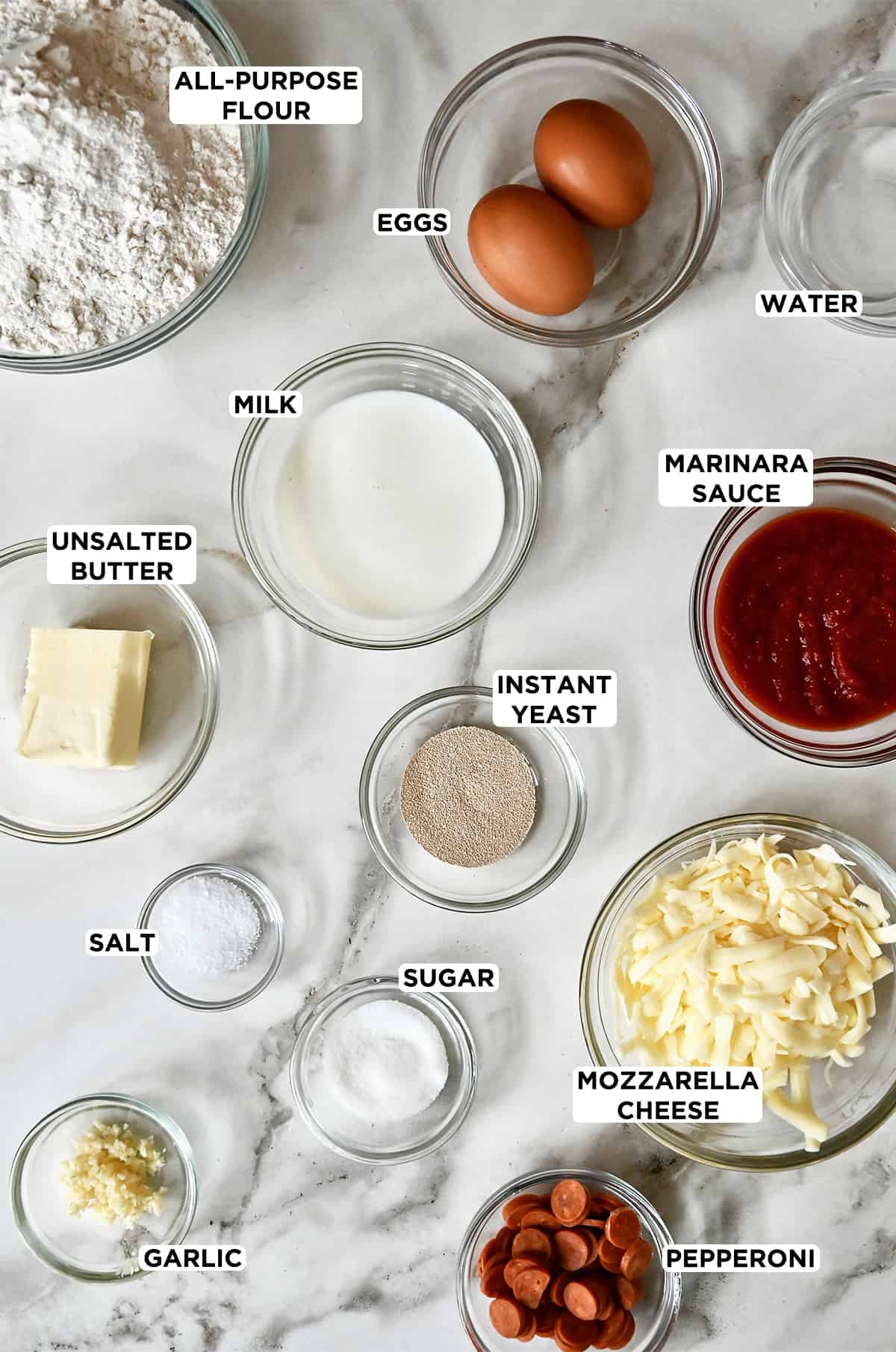 Various sizes of clear bowls containing flour, eggs, milk, marinara sauce, instant yeast, shredded mozzarella cheese, salt, sugar, butter and mini pepperoni.