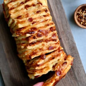 A hand reaches for a pepperoni pull-apart pizza bread on a wood cutting board.