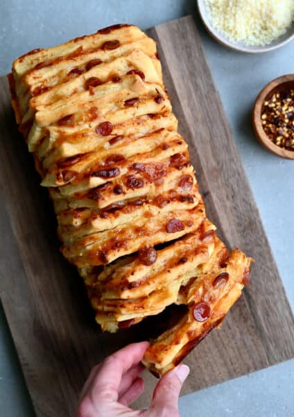A hand reaches for a pepperoni pull-apart pizza bread on a wood cutting board.