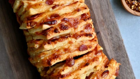 A hand reaches for a pepperoni pull-apart pizza bread on a wood cutting board.