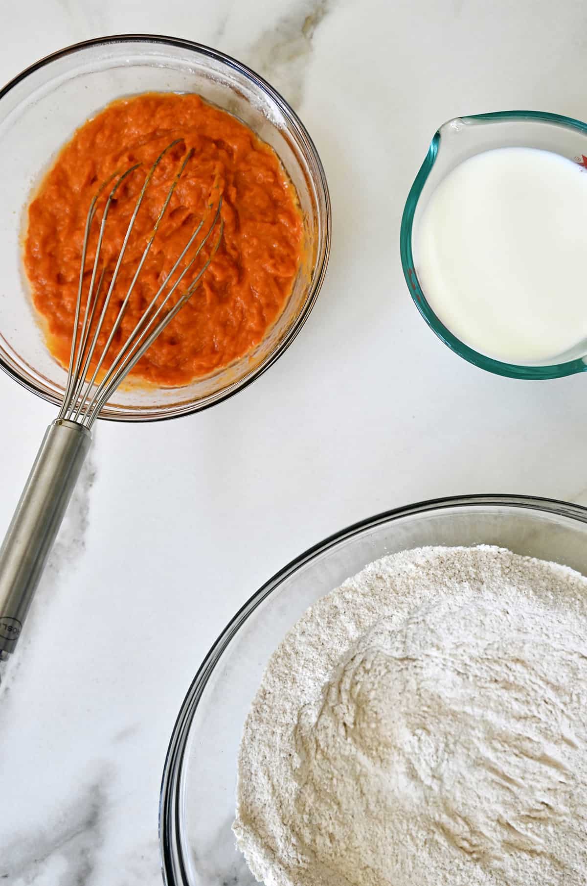 A clear bowl containing all-purpose flour next to a clear bowl containing pumpkin puree and a whisk next to a liquid measuring cup containing milk.