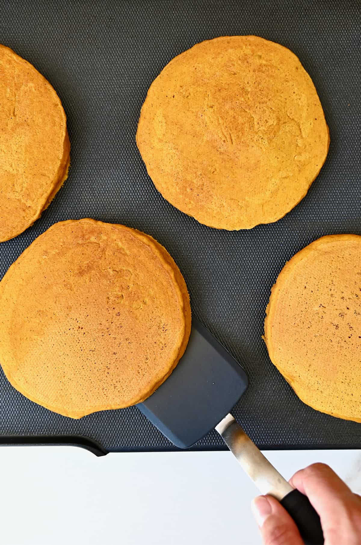 A hand holds a spatula under a pumpkin pie pancake on a griddle next to three other pancakes.