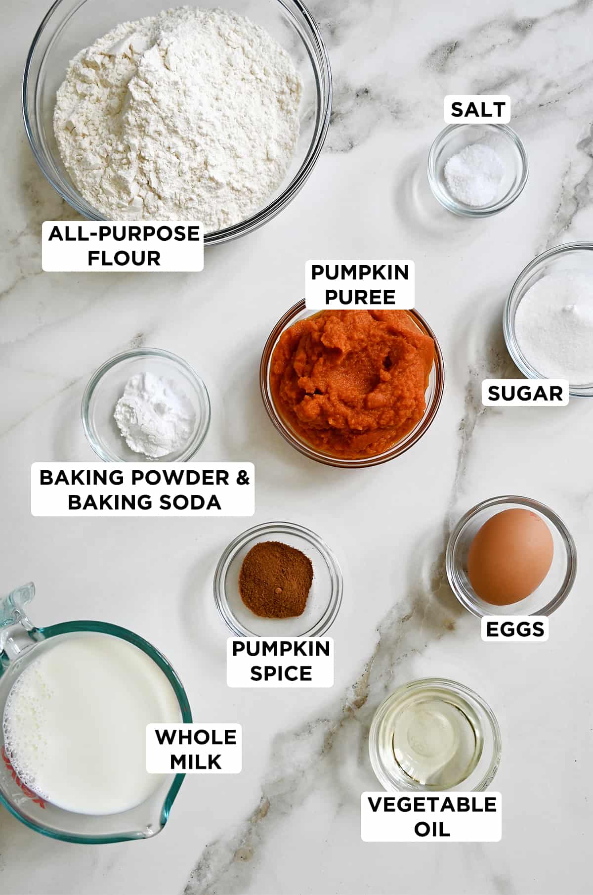 Various sizes of clear bowls containing flour, pumpkin puree, an egg, sugar, baking powder and baking soda, pumpkin spice, salt, oil and whole milk.