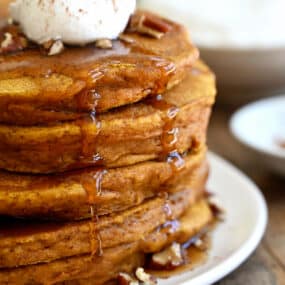 A tall stack of pumpkin pie pancakes topped with pecans, whipped cream and maple syrup.