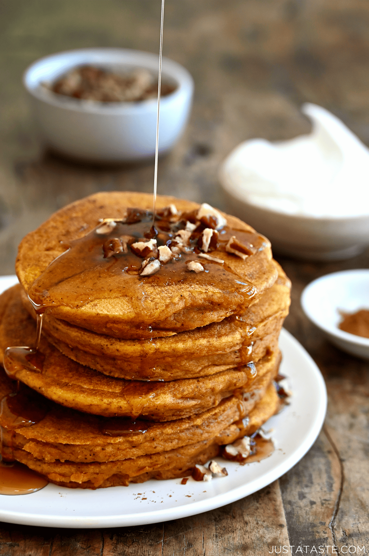 A tall stack of pumpkin pancakes topped with pecans and drizzled with maple syrup on a white plate.