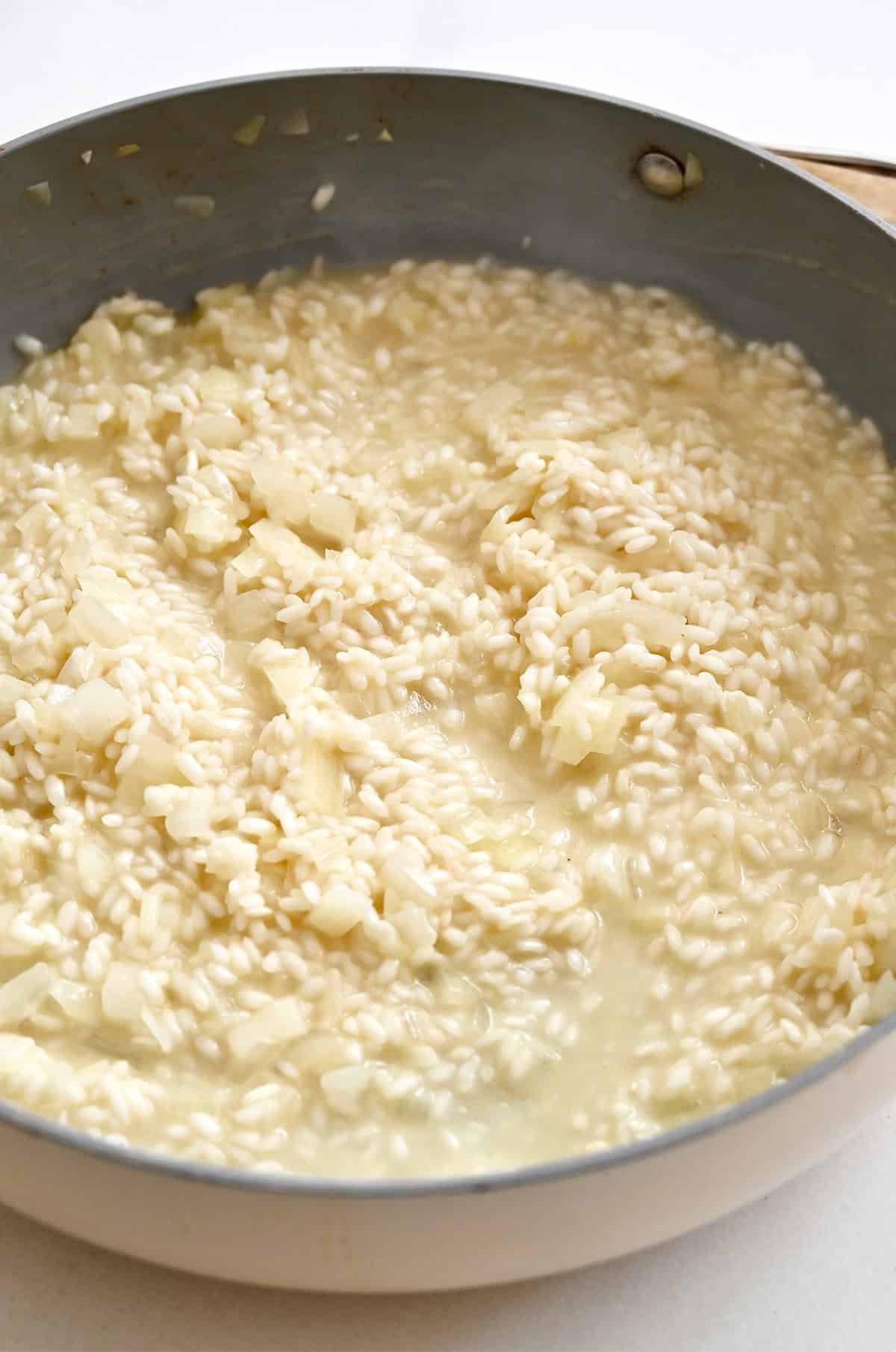 Arborio rice and diced onion cooking in a large skillet after the white wine has been absorbed, showing the creamy risotto base.