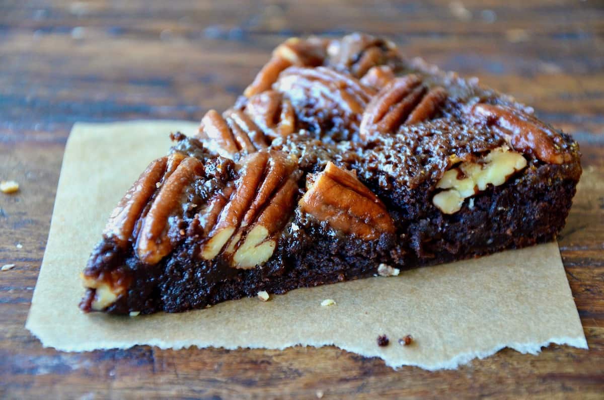 A wedge-shaped slice of Pecan Pie Brownie sits on a scrap of brown parchment paper on a wood surface.