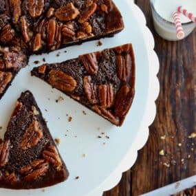 Slices of Pecan Pie Brownies sit on a white scalloped cake stand. Nearby is a glass of milk with two striped straws.