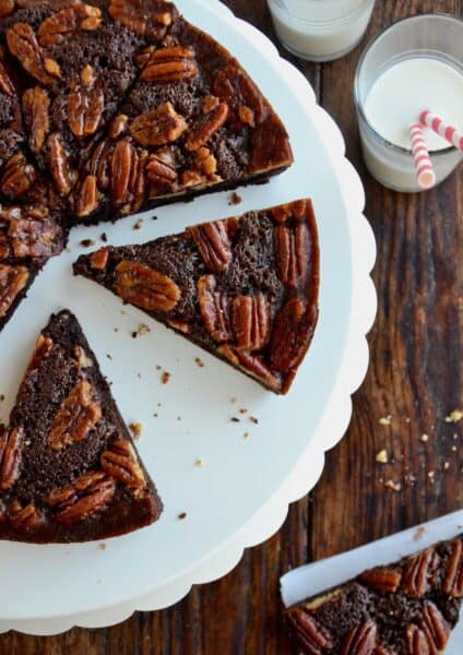 Slices of Pecan Pie Brownies sit on a white scalloped cake stand. Nearby is a glass of milk with two striped straws.