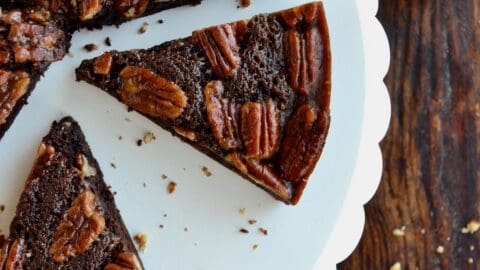 Slices of Pecan Pie Brownies sit on a white scalloped cake stand. Nearby is a glass of milk with two striped straws.