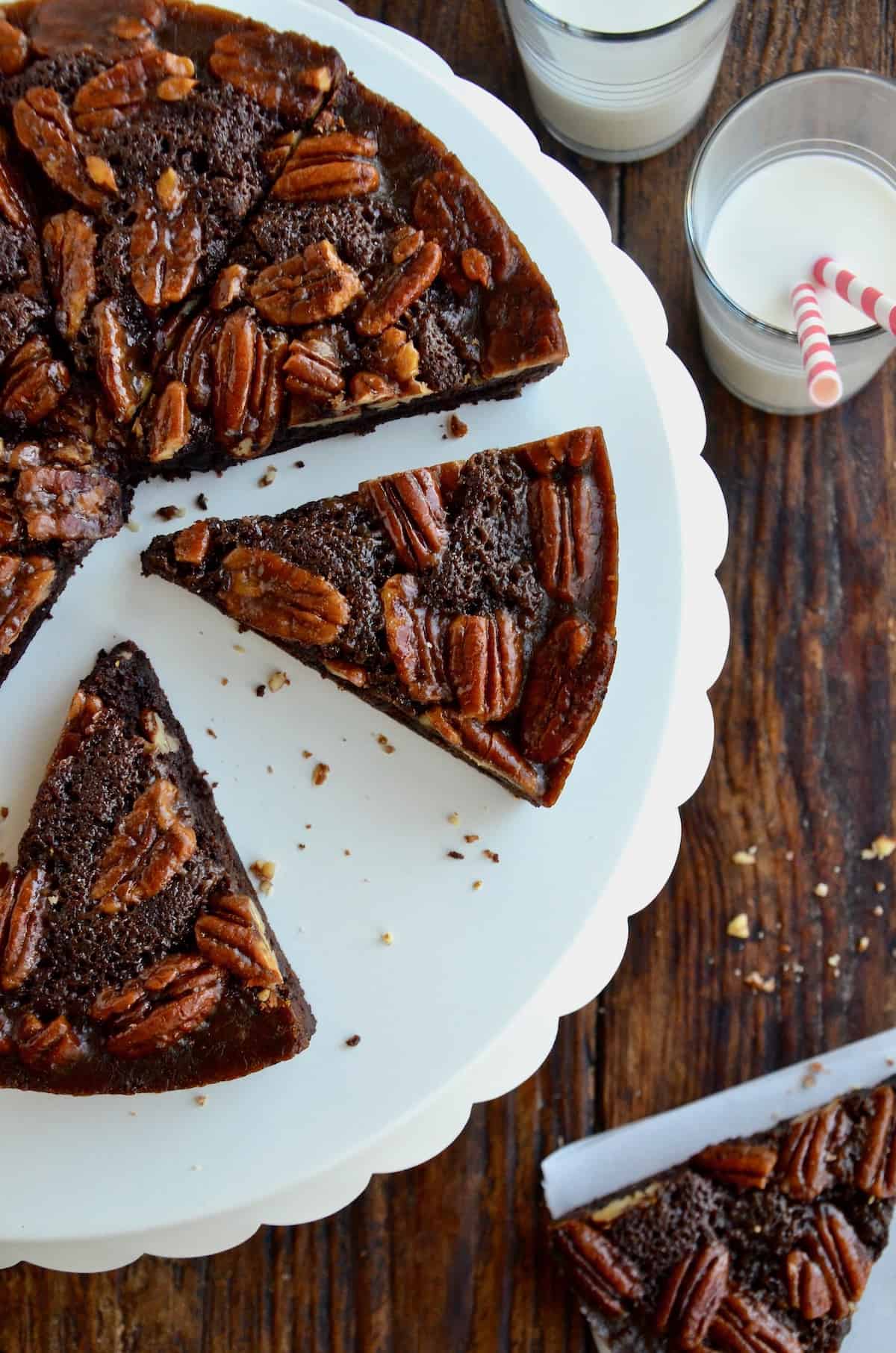 Slices of Pecan Pie Brownies sit on a white scalloped cake stand. Nearby is a glass of milk with two striped straws.