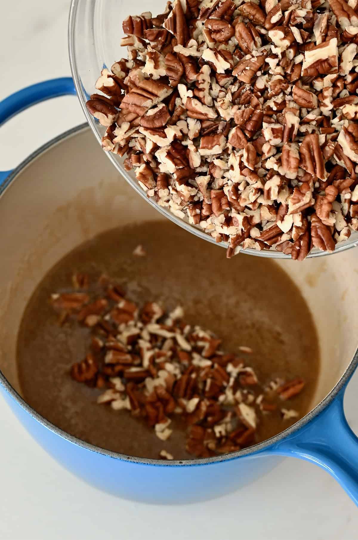 Adding chopped pecans to a caramel-like mixture made with honey, brown sugar and heavy cream in a saucepan.