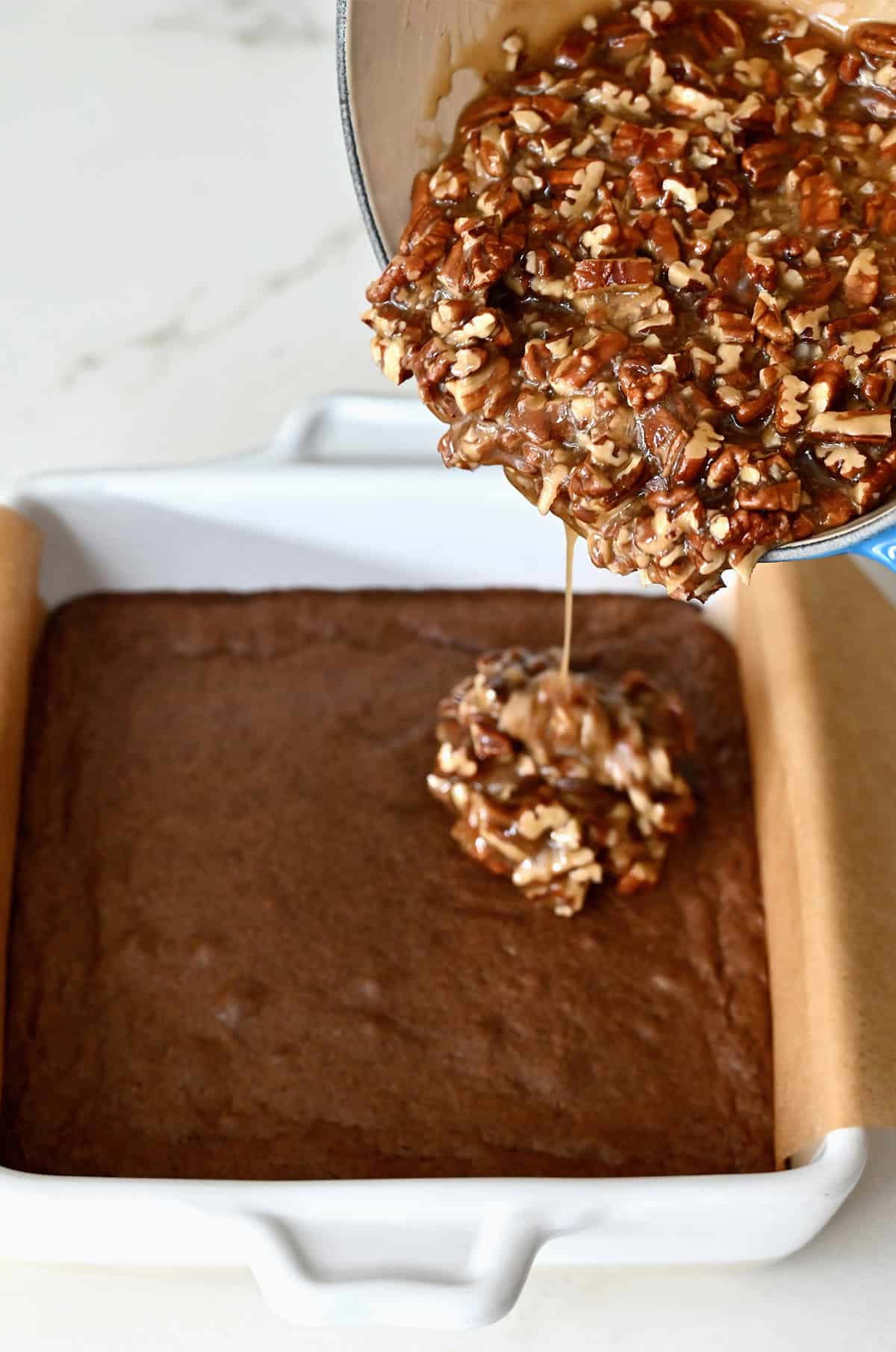 Pouring a honey-sweetened pecan pie filling atop par-baked brownies in a square pan.