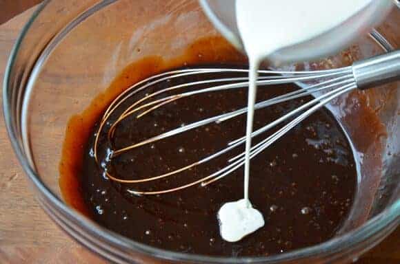 Milk being poured into a bowl containing chocolate molasses cake batter and a whisk.