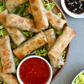 Crispy baked chicken spring rolls on a bed of shredded green cabbage next to a small bowl containing sweet chili sauce.