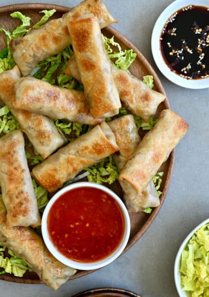 Crispy baked chicken spring rolls on a bed of shredded green cabbage next to a small bowl containing sweet chili sauce.
