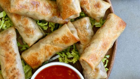 Crispy baked chicken spring rolls on a bed of shredded green cabbage next to a small bowl containing sweet chili sauce.