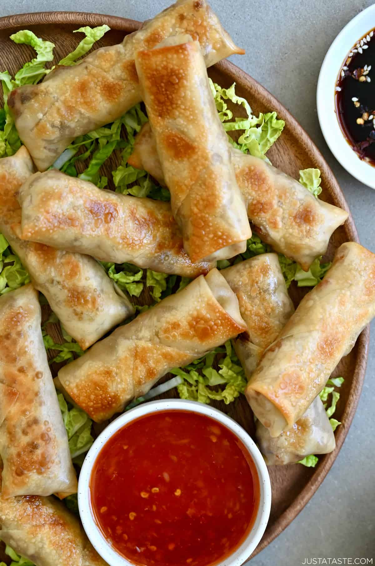 Golden brown baked chicken spring rolls atop a bed of shredded green cabbage next to a small bowl containing Thai sweet chili sauce.