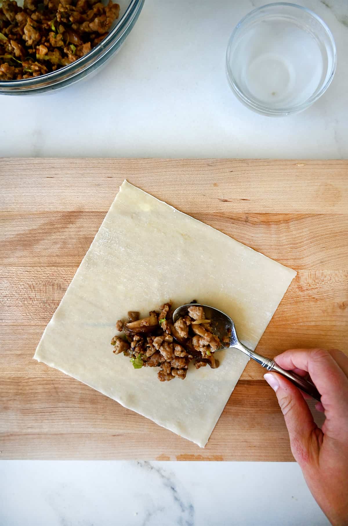 A hand holding a spoon adds a cooked chicken mixture atop a spring roll wrapper. 