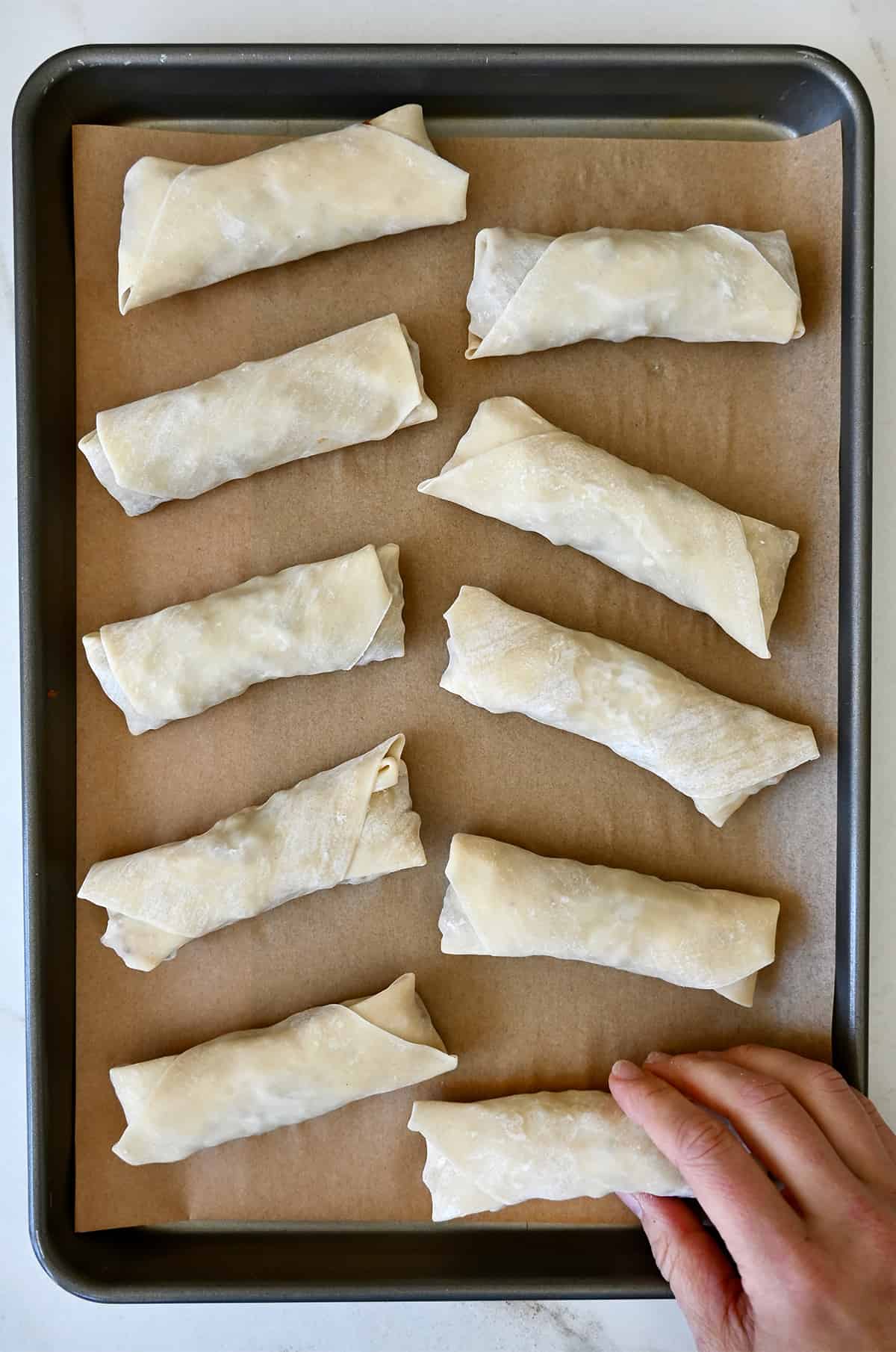 Ten unbaked chicken spring rolls on a parchment paper-lined baking sheet.