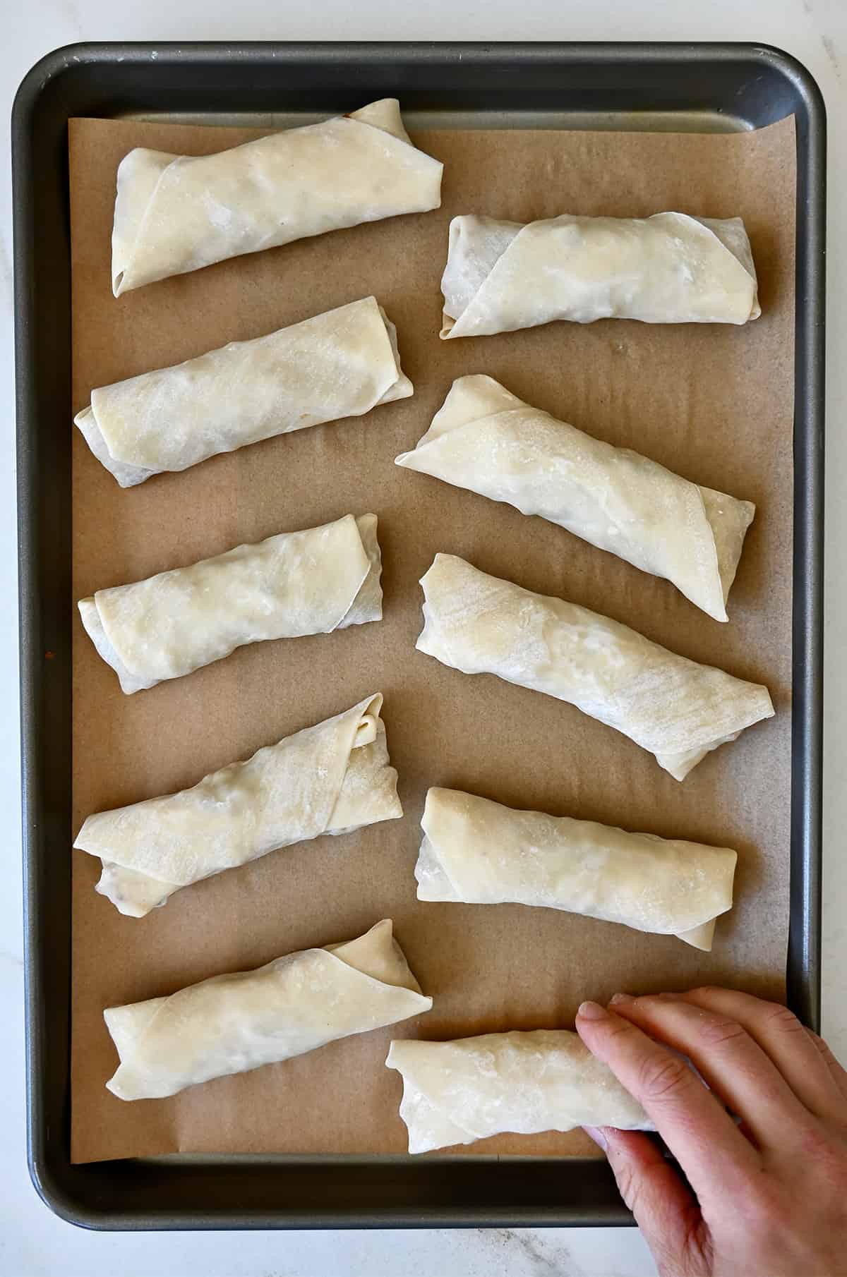 Ten unbaked chicken spring rolls on a parchment paper-lined baking sheet.