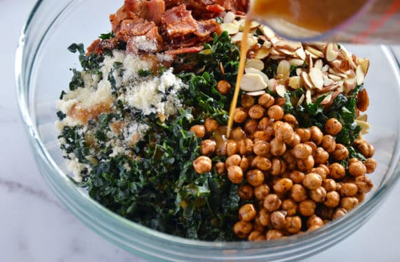 Warm bacon dressing being poured onto a glass bowl containing kale salad ingredients