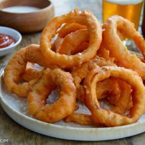 A plate of onion rings with Ranch dressing, ketchup and beer in the background