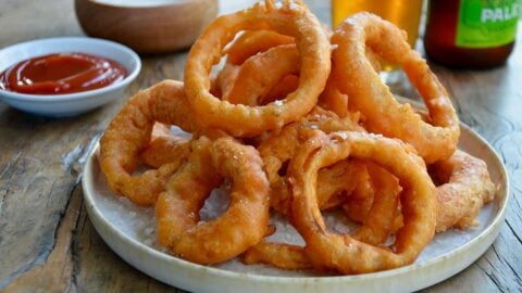 A plate of onion rings with Ranch dressing, ketchup and beer in the background