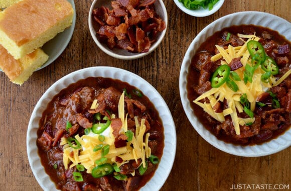 Top-down view of bowls of homemade Chili con Carne topped with, sour cream, shredded cheddar cheese and jalapeños next to a small bowl containing pieces of bacon and a plate with cornbread 