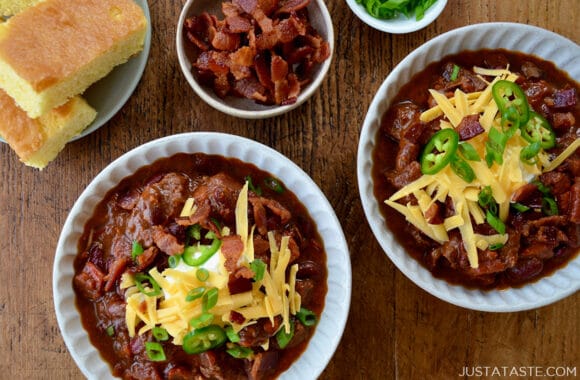 Top-down view of bowls of homemade Chili con Carne topped with, sour cream, shredded cheddar cheese and jalapeños next to a small bowl containing pieces of bacon and a plate with cornbread