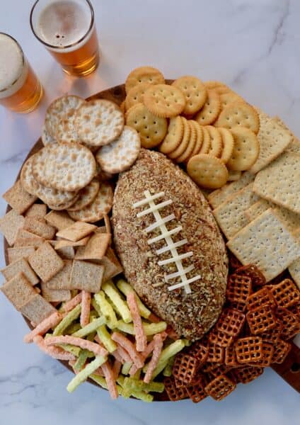 A football-shaped cheeseball on a charcuterie board surrounded by different types of crackers, veggie straws and pretzels.