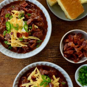 Top-down view of bowls of Chili con Carne topped with, sour cream, shredded cheese and jalapeños next to a small bowl containing pieces of bacon and a plate with cornbread