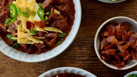 Top-down view of bowls of Chili con Carne topped with, sour cream, shredded cheese and jalapeños next to a small bowl containing pieces of bacon and a plate with cornbread