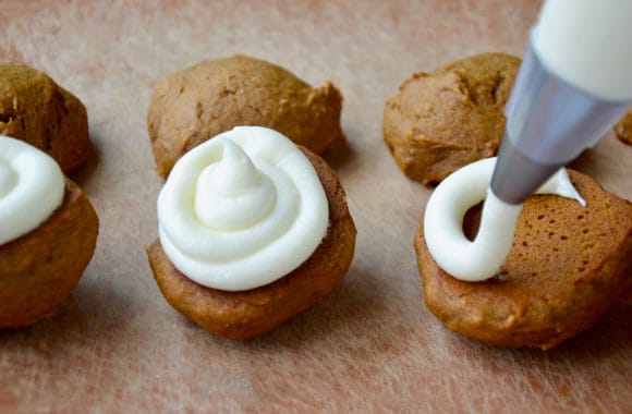 Pumpkin whoopie pies on a wood board being filled with cream cheese frosting in a piping bag