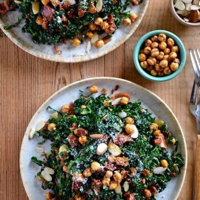 Two white plates piled high with kale salad with a small bowl of crispy chickpeas next to it