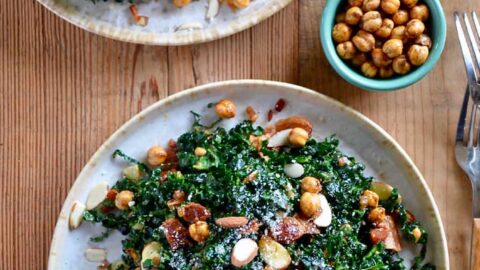 Two white plates piled high with kale salad with a small bowl of crispy chickpeas next to it