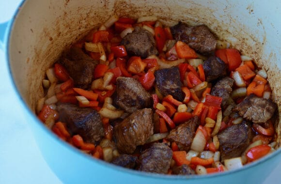 Large blue stockpot containing seared meat, red bell peppers and onions