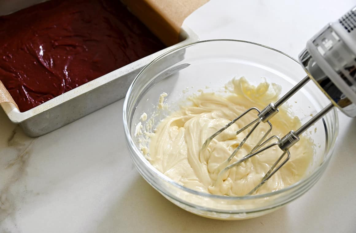 A hand beater in a clear bowl containing cheesecake mixture