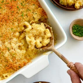 A hand holding a spoon in a baking dish of Roasted Garlic Macaroni and Cheese