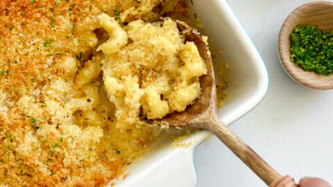 A hand holding a spoon in a baking dish of Roasted Garlic Macaroni and Cheese