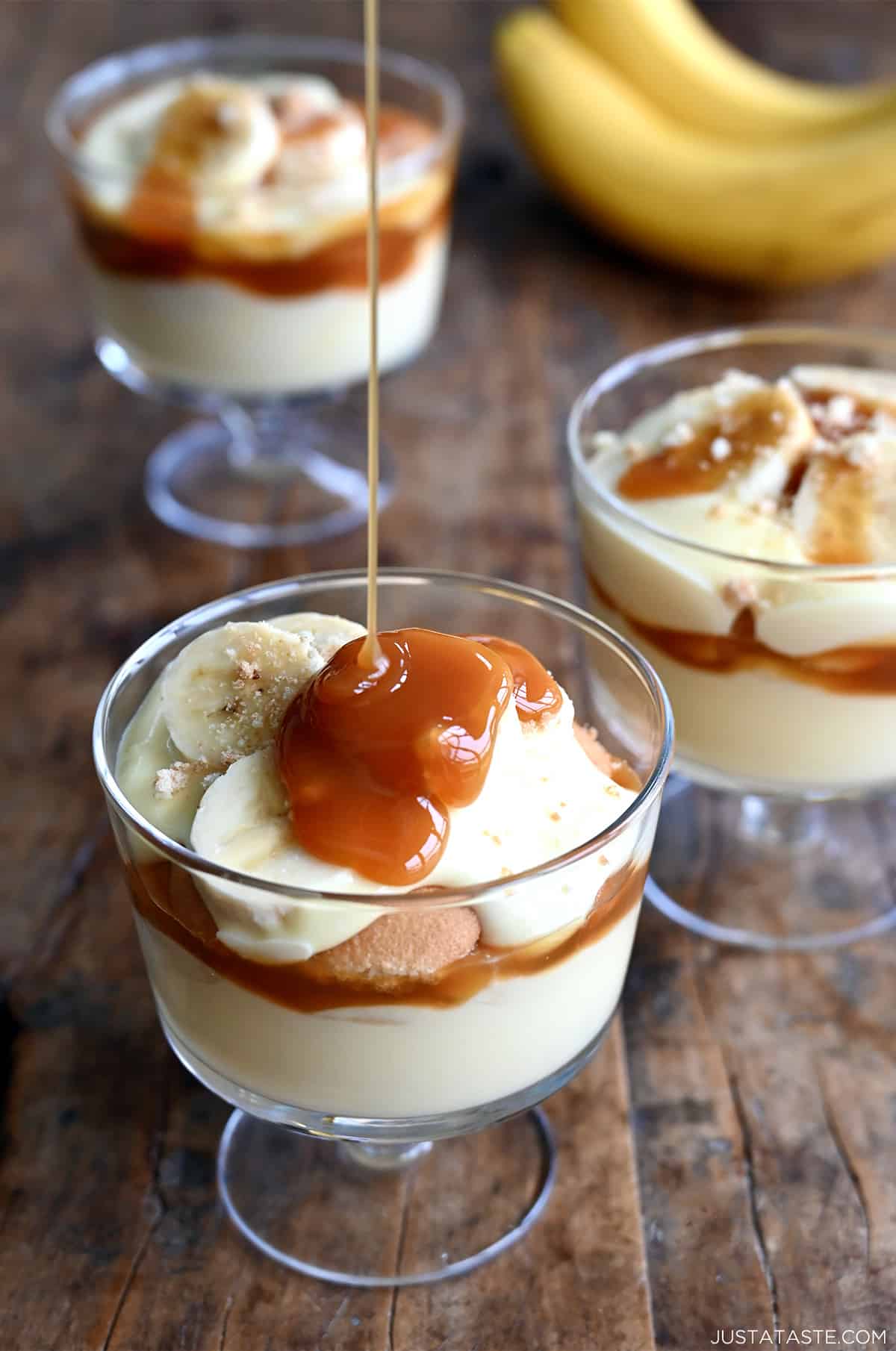 Salted caramel sauce being drizzled atop sliced bananas in a parfait glass with layers of banana pudding and vanilla wafers.