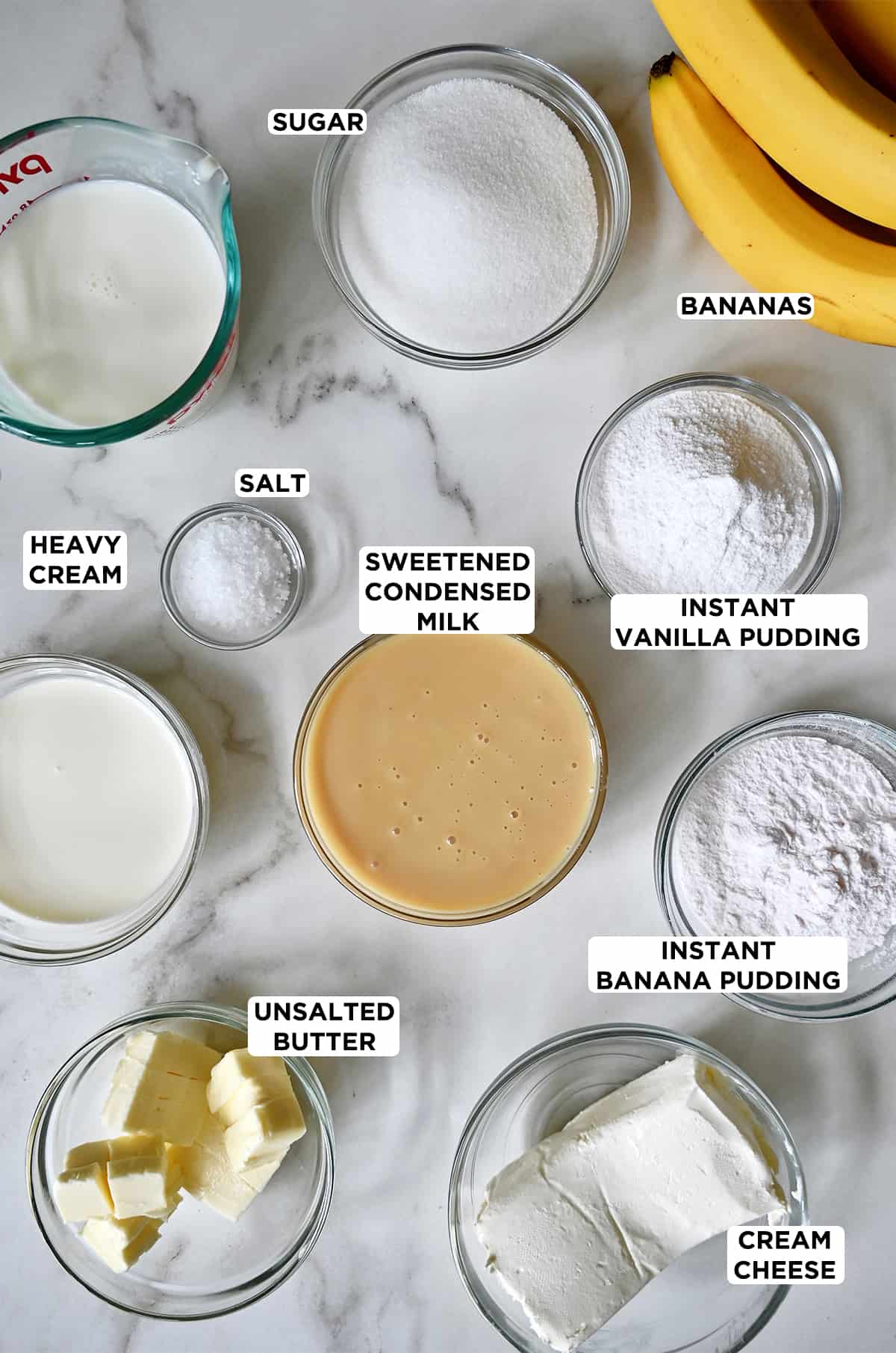 Various sizes of glass bowls containing banana pudding and salted caramel ingredients, including heavy cream, sugar, instant vanilla and banana puddings, salt, sweetened condensed milk, butter, cream cheese and whole milk. Two bananas are next to the bowls.